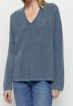 Woman wearing a blue V-neck long-sleeve sweater paired with blue jeans, standing against a plain light background.