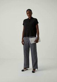 Camicia nera a maniche corte con bottoni, pantaloni grigi a gamba larga, scarpe bianco e nere, in mano una borsa a tracolla grigia chiara, in piedi su uno sfondo neutro.
