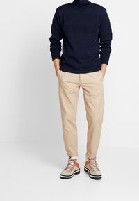 Maglione a collo alto lavorato a maglia di colore blu navy con polsini a costine, abbinato a pantaloni beige e stivali da escursionismo multicolori con dettagli arancioni e lacci.