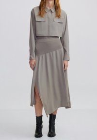 Eine Frau trägt eine taubfarbene, knöchellange, mit Knöpfen versehene Jacke und einen hoch taillierten, asymmetrischen Rock mit seitlichem Schlitz, kombiniert mit schwarzen Knöchelstiefeln.