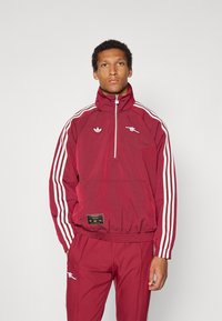 Casaco corta-vento vermelho com riscas brancas da Adidas, gola alta, bolso frontal e logótipo do Arsenal FC; combinado com calças vermelhas a condizer.