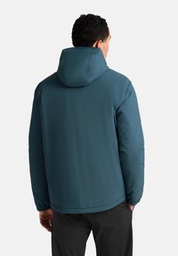 Giacca isolante color turchese con cappuccio, dotata di polsini elasticizzati e una texture liscia. La parte posteriore presenta un design semplice senza decorazioni.