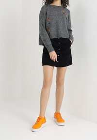 Femme portant un pull gris avec des broderies colorées, une jupe noire à boutons et des sneakers orange vif, se tenant avec une main dans la poche.
