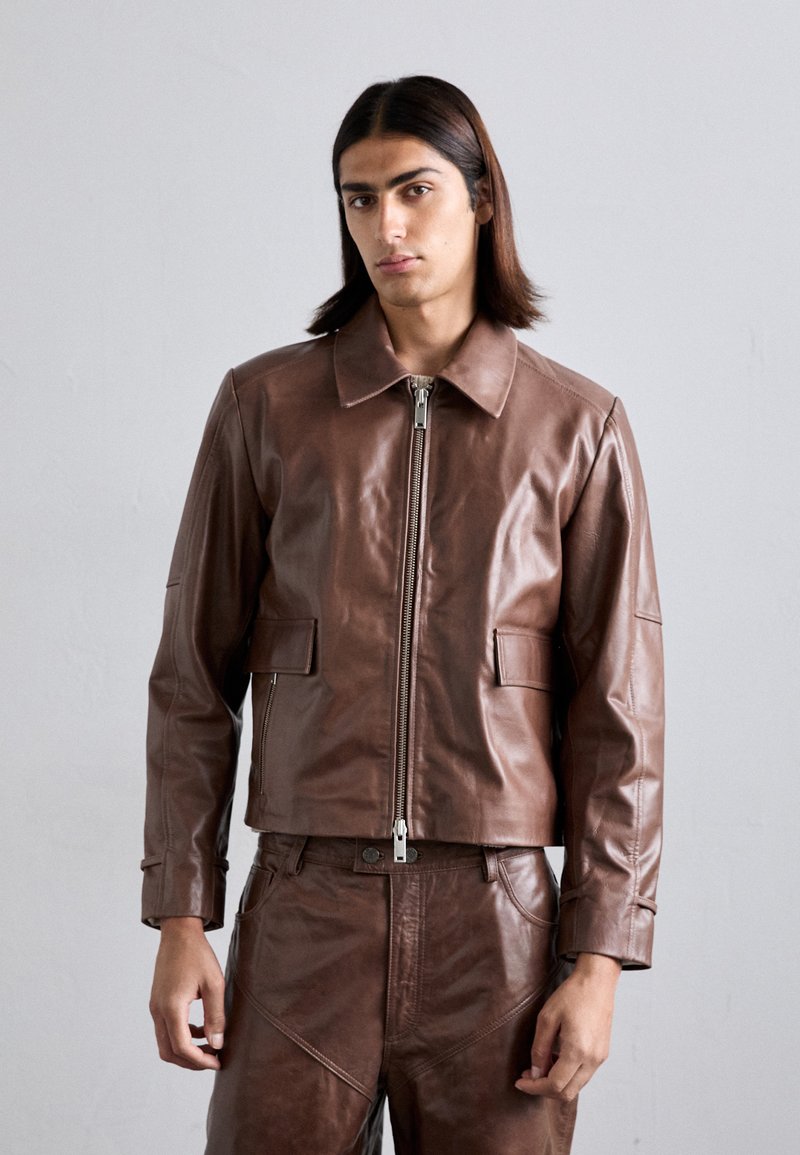 Giacca in pelle marrone con collo, chiusura frontale a zip e due tasche quadrate, caratterizzata da una texture liscia e una vestibilità su misura.