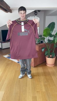 Tricou bordeaux cu accente galbene și logo Puma, cu mâneci scurte, guler rotund, ținut de o persoană într-un living cu plante și mobilă.