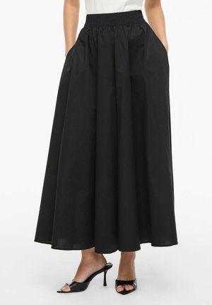 Mustast pikast A-lõikeline seelik, millel on elastne vöökoht ja küljetaskud, kantud mustade avatud varvastega kontsakingade ja valge topiga.