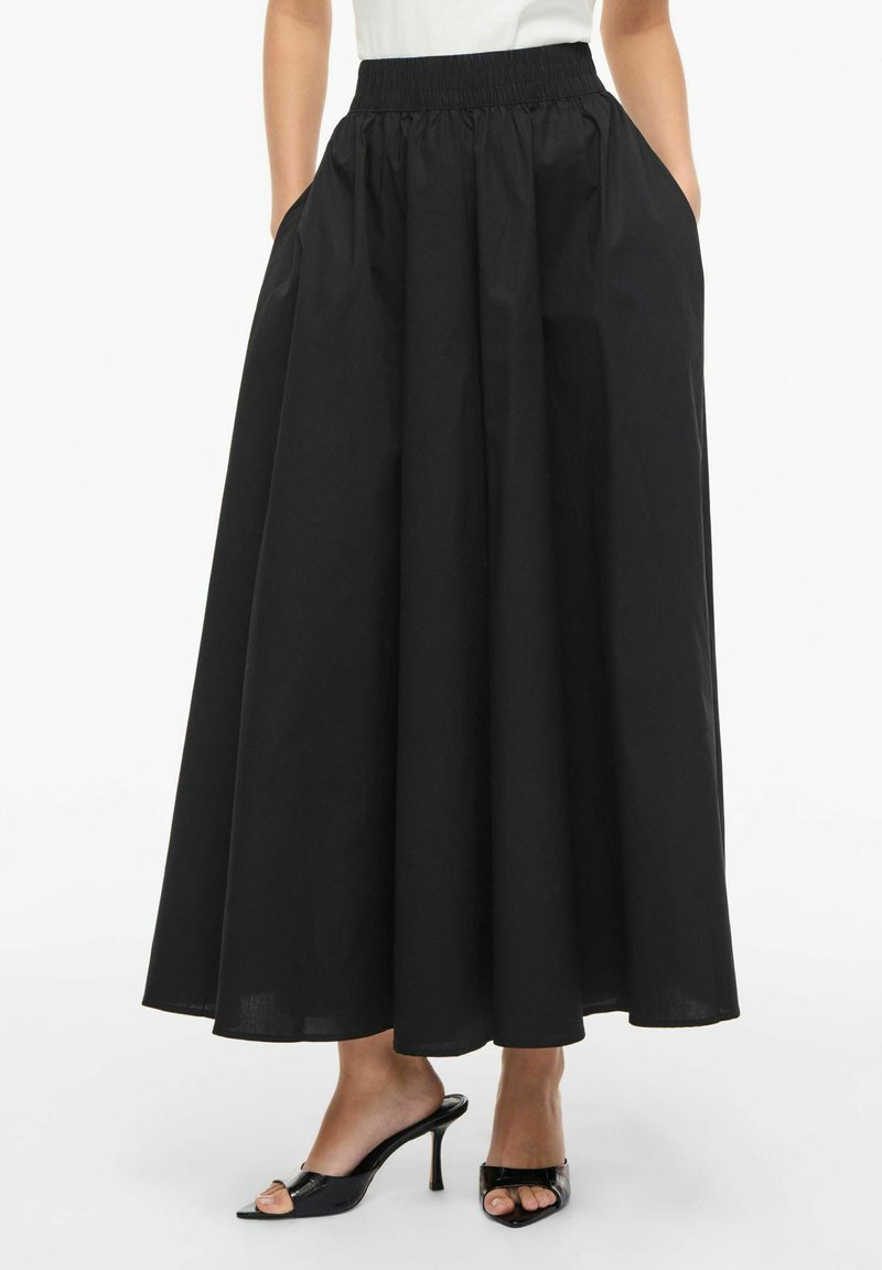 Črna dolga trapezasta krilo z elastičnim pasom in stranskimi žepi, nošena s črnimi odprtimi natikači z visoko peto in belim zgornjim delom.