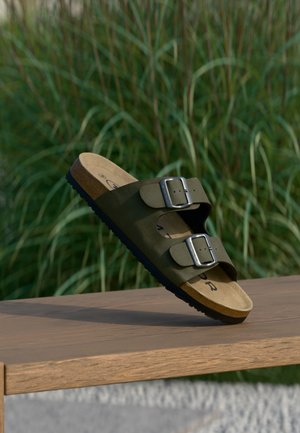 Grüne Wildledersandale mit zwei verstellbaren silbernen Schnallen, Korkfußbett und einer strukturierten schwarzen Gummisohle, auf einer Holzoberfläche präsentiert.