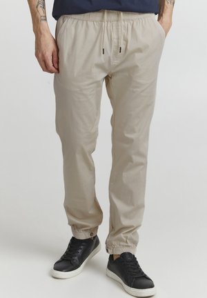 Beige, lette joggerbukser med elastisk talje og manchetter, der har to sidelommer. Bæres med sorte sneakers.