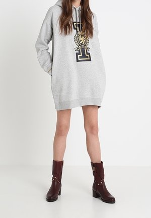 Femme portant une robe sweat-shirt gris clair oversize avec un grand emblème sur le devant et des bottes à talons brun foncé mi-mollet, debout devant un fond blanc.