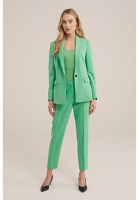 Zelený elegantní blejzr a kalhoty ve stejném odstínu, vyrobené z hladké látky. Blejzr má jeden knoflík a klopové límce. Černé špičaté podpatky.