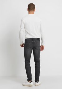 Pantalones vaqueros negros ajustados con una textura sutil, que presentan un diseño clásico de cinco bolsillos y un pequeño parche con logo en la parte trasera. Combinados con una camiseta blanca.