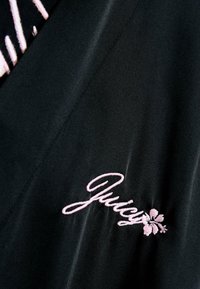 Melna auduma ar izšuvumu vārdu "Juicy" rozā kursīvā ar ziedu akcentu. Gluda tekstūra ar nedaudz spīdīgu apdari.