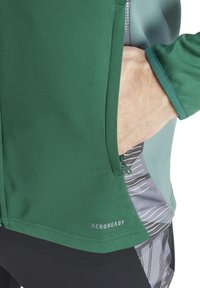 Top sportivo verde con una texture liscia, dotato di una tasca con zip, pannelli laterali a motivo grigio e branding "AEROREADY" vicino all'orlo.