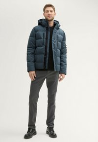 Modra napihnjena jakna s kapuco, ki vključuje zapiranje z zadrgo in gumbi ter stranska žepa. V kombinaciji s sivimi hlačami in črnimi čevlji.