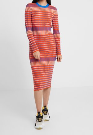 Femme portant une robe rayée multicolore longueur genoux à manches longues, aux tons rouge, violet et pêche, associée à des baskets blanches et jaunes.