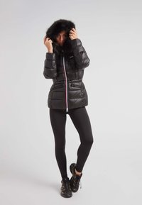 Czarna kurtka puchowa z kapturem wykończonym sztucznym futrem, zapięciem na suwak z przodu, elastycznym pasem oraz bocznymi kieszeniami na suwak. Noszona z czarnymi legginsami i butami.