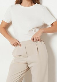 T-shirt blanc à manches courtes associé à un pantalon beige plissé taille haute, avec une texture lisse et une silhouette ajustée.