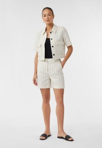 Kurzärmeliger, gestreifter Blazer und passende Shorts in Creme mit schwarzen Linien. Der Blazer hat eine Tasche und schwarze Knöpfe; getragen mit einem schwarzen Oberteil und Sandalen.