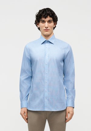 Άνδρας που φοράει ένα ανοιχτό μπλε πουκάμισο με κατακόρυφες ρίγες και μακριά μανίκια και μπεζ παντελόνι, στέκεται μπροστά σε απλό φόντο.