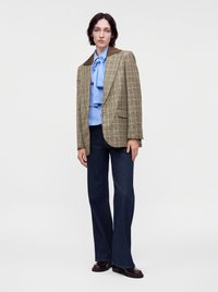 Femme debout portant un blazer marron à carreaux sur un chemisier bleu avec un nœud, un pantalon évasé sombre et des chaussures foncées sur un fond blanc uni.