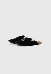Pantuflas de terciopelo negro con detalle del logo en oro, abiertas por detrás y forradas con un estampado de leopardo en el interior. Textura suave y forma redondeada para mayor comodidad.
