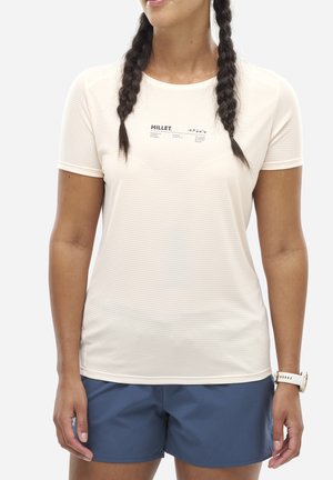 Femme aux cheveux tressés portant un t-shirt blanc Millet, un short bleu et une montre connectée blanche, debout avec les bras détendus le long du corps.