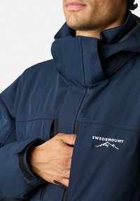 Navy blue jacket featuring a high collar, structured design, and logo detail. Includes black accents and a velcro cuff closure.