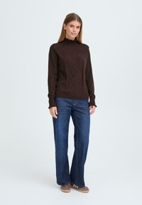 Brun strikket genser med høy krage og diamantmønster foran, sammen med vide denimjeans og brune sko.