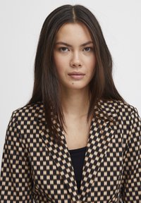 Brauner und schwarzer karierter Blazer mit glatter Textur, ausgestattet mit eingeschliffenen Revers und einer taillierten Passform. getragen über einem schwarzen Oberteil.