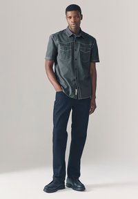 Camicia scura a manica corta con bottoni e due tasche sul petto, cuciture a contrasto; abbinata a pantaloni blu navy e scarpe nere, in posizione eretta.