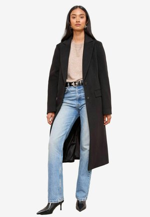Zwarte lange jas over een beige top, lichtblauwe high-waisted jeans en zwarte puntige hakken. Gladde stof met een getailleerde pasvorm en geknipte revers.