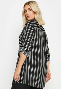 Camicia a righe bianco e nere con maniche arrotolate. Realizzata in tessuto leggero, presenta spacchi laterali e una vestibilità comoda. Dettagli sul retro visibili.