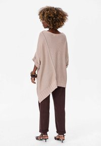 Beige gebreide poncho met oversized mouwen en een zijsplit. Gecombineerd met donkerbruine broeken en gespsandalen; heeft een gestructureerde stof.