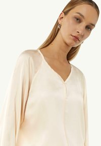 Helles Creme, V-Ausschnitt-Bluse aus Satin mit langen Ärmeln, weicher Textur und subtiler glanz, mit einem Knopfdetail entlang der Vorderseite.
