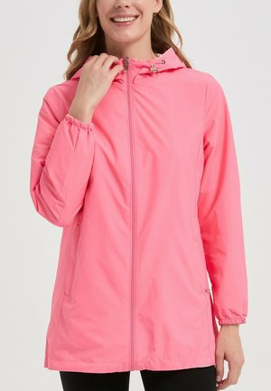 Femme portant une veste à capuche rose vif avec des poignets élastiques, une fermeture éclair frontale et des poches latérales zippées, souriant et touchant le col zippé.