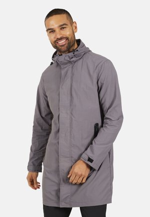 Homme souriant portant une veste à capuche gris clair avec fermeture éclair à l'avant et bandes auto-agrippantes aux poignets, debout devant un fond uni.