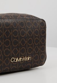 Sac marron Calvin Klein avec un motif monogramme "ck" en répétition et un logo Calvin Klein doré sur une surface texturée, sur un fond clair.