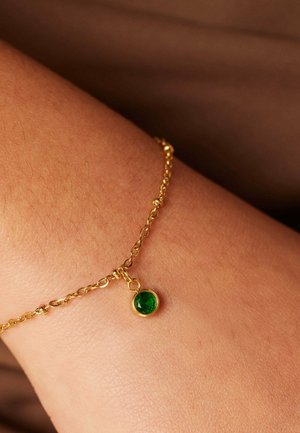 Gold chain bracelet with a small green faceted stone pendant. The bracelet is worn on a wrist against skin, showcasing its shiny texture.