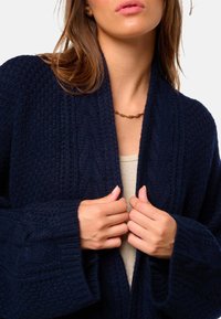 Cardigan en maille bleu marine avec un motif texturé, manches larges et ouverture sur le devant, associé à un haut de couleur claire et un collier en or.