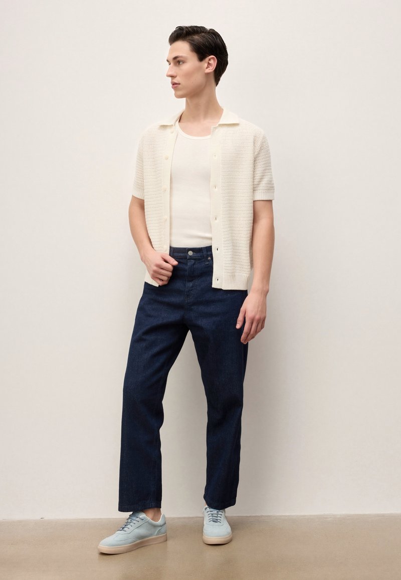 Jeune homme debout, portant une chemise blanche à manches courtes boutonnée sur un débardeur blanc, un jean bleu foncé et des baskets bleu clair.