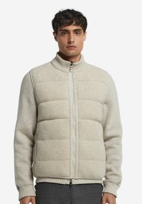 Beige Strickjacke mit einem Reißverschluss auf der Vorderseite, gerippter Textur an Schultern und Hals und gepolsterten, gesteppte Abschnitten am Körper. Normale Passform, hoher Kragen.