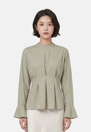 Vrouw met kort donker haar, die een lichtgroene longsleeve top draagt met geplisseerde taille en uitlopende manchetten, staand tegen een effen achtergrond.