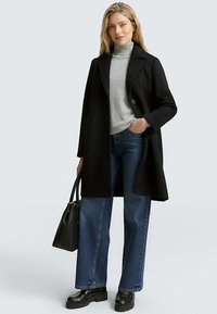 Czarny płaszcz z mieszanki wełny z kołnierzem z wycięciem, zapięciem na jeden guzik, klapkami na ramionach, zestawiony z szarym golfem i szerokimi niebieskimi dżinsami.