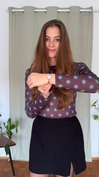 Strieborné náramkové hodinky so štvorcovým ciferníkom, nosené na zápästí; dlhý rukáv topu s fialovými bodkami a čierna sukňa; vnútorné prostredie.