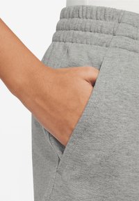 Šedé bavlněné tepláky s žebrovaným elastickým pasem a boční kapsou, na detailním záběru je vidět ruka opřená v kapse.