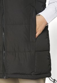 Svart vadderad väst med frontdragkedja, sidofickor och justerbar nederkant. Materialet verkar vara slätt med en matt yta.