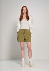 Pantaloni corto verde oliva con vita elasticizzata, tasche frontali e orli arrotolati, abbinati a un lungo cardigan bianco e sneakers bianco sporco.