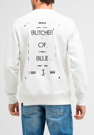 Cremfarbener Sweatshirt mit strukturierter Oberfläche, von hinten mit schwarzen Drucken, einschließlich der Worte "BUTCHER OF BLUE". Sichtbare blaue Jeans.
