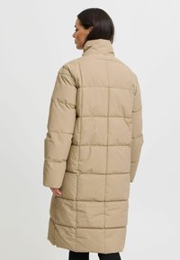 Abrigo de plumas beige con un cuello alto y diseño de acolchado en patchwork. Presenta mangas largas y un corte a media longitud. Textura de tela suave.